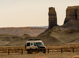 An RV rental camper van parked in a vast, arid landscape with impressive desert rock formations and a wooden fence.