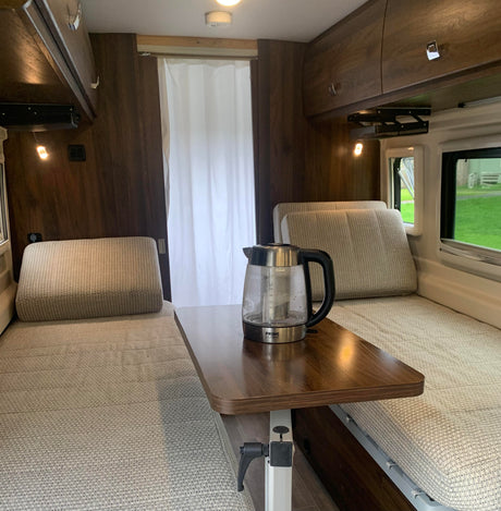 RV rental interior features light fabric bench seating/beds, a central adjustable table with a kettle, and overhead cabinets.