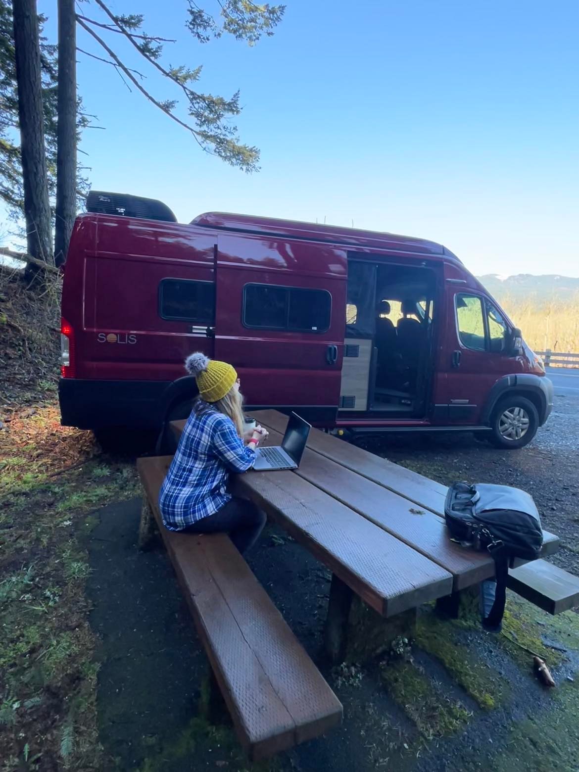 Woman works on laptop at picnic table beside a red Solis camper van. Remote office ideal with RV rental in nature.