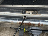 Close-up of an RV rental's rear. Features rusty chrome bumper, hitch wiring, and a jack support rod entering the body.