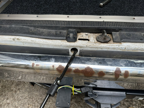 Close-up of an RV rental's rear. Features rusty chrome bumper, hitch wiring, and a jack support rod entering the body.