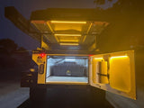 Illuminated overland RV rental: a rugged camper with a rooftop tent and bright LED-lit gear storage, ready for off-grid adventures.