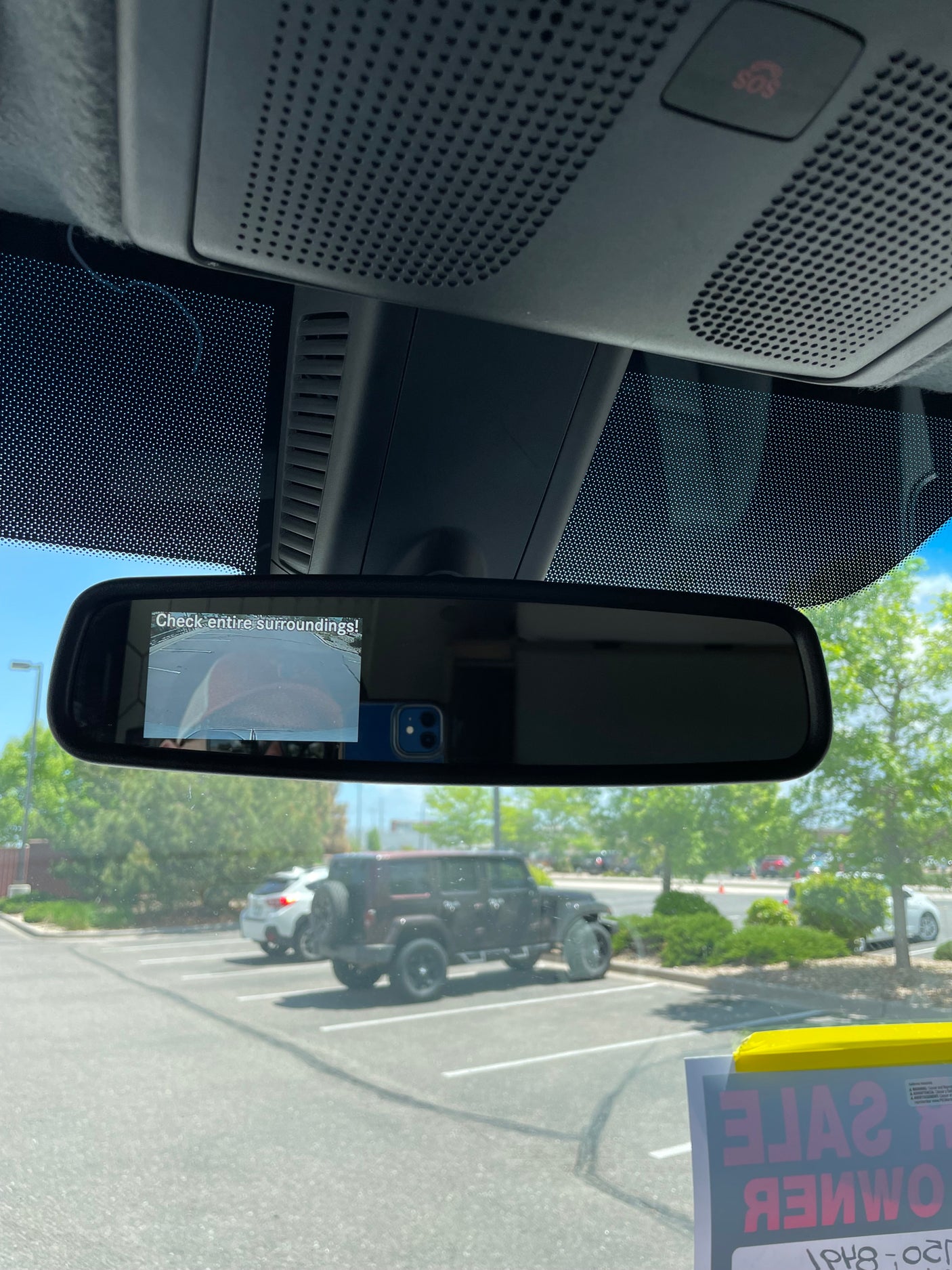 Inside a vehicle: rearview mirror with backup camera. Outside, a parking lot with vehicles, incl. a 'FOR SALE' SUV. Great for rv rental ideas.