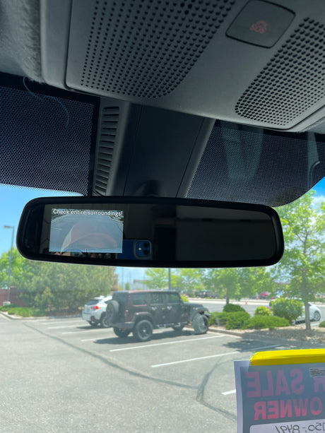 Inside a vehicle: rearview mirror with backup camera. Outside, a parking lot with vehicles, incl. a 'FOR SALE' SUV. Great for rv rental ideas.