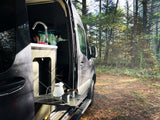 This RV rental offers a perfect nature escape! A camper van with an open side door reveals a sink & pull-out table with coffee setup, surrounded by forest.