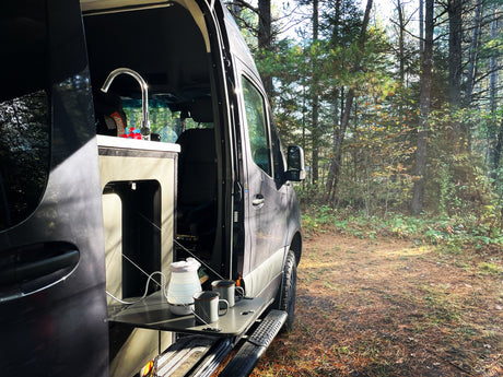 This RV rental offers a perfect nature escape! A camper van with an open side door reveals a sink & pull-out table with coffee setup, surrounded by forest.