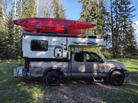 Adventure awaits! Gray pickup truck with Palomino truck camper & red kayaks on top, parked in woods. Ideal for rv rental.