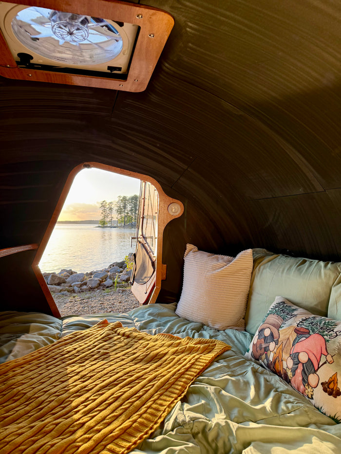 Inside a cozy RV rental, a comfy bed faces an open door with a beautiful lake & sunset view. Ideal for camping.