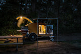 An illuminated yellow teardrop RV rental camper at night, featuring a cozy interior, open cooking area, and picnic setup.