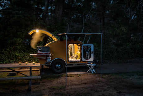 An illuminated yellow teardrop RV rental camper at night, featuring a cozy interior, open cooking area, and picnic setup.