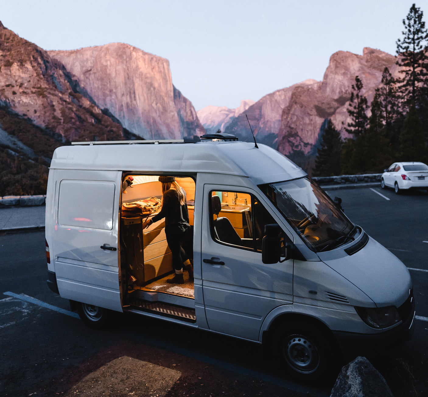 Person inside a cozy, illuminated RV rental camper van, parked amidst iconic Yosemite Valley mountains at dusk.