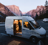 Person inside a cozy, illuminated RV rental camper van, parked amidst iconic Yosemite Valley mountains at dusk.