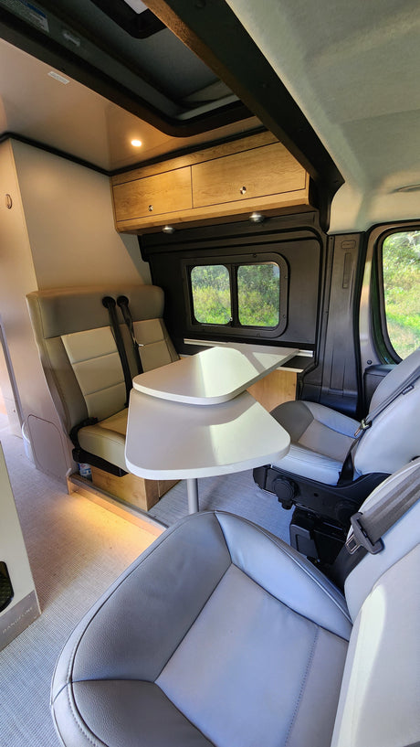 Spacious RV rental interior with light-toned seating, a curved dining table, overhead cabinets, and large windows with scenic views.