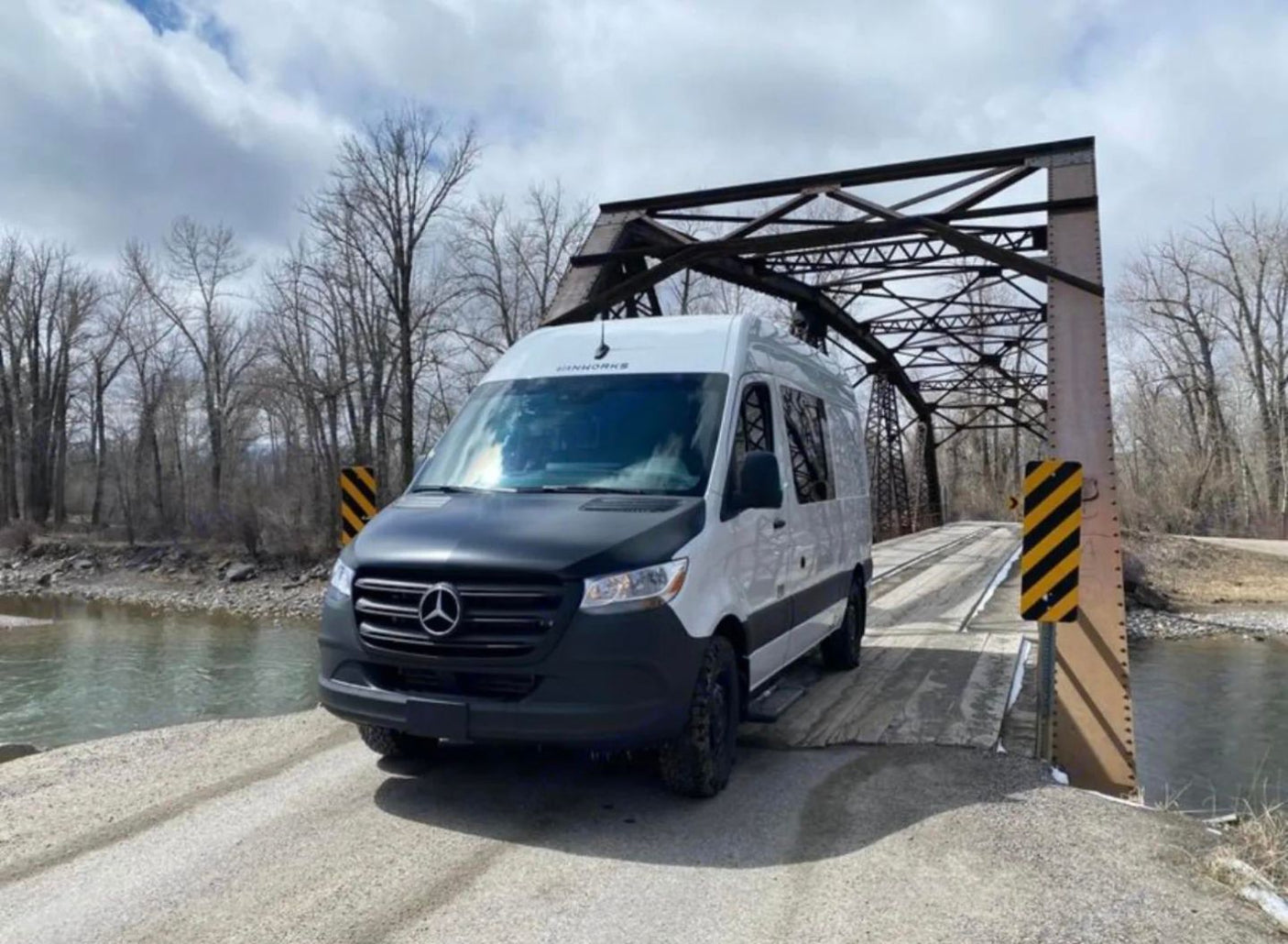 White Sprinter van RV rental on a rustic wooden bridge over a river with bare trees. Ready for adventure!