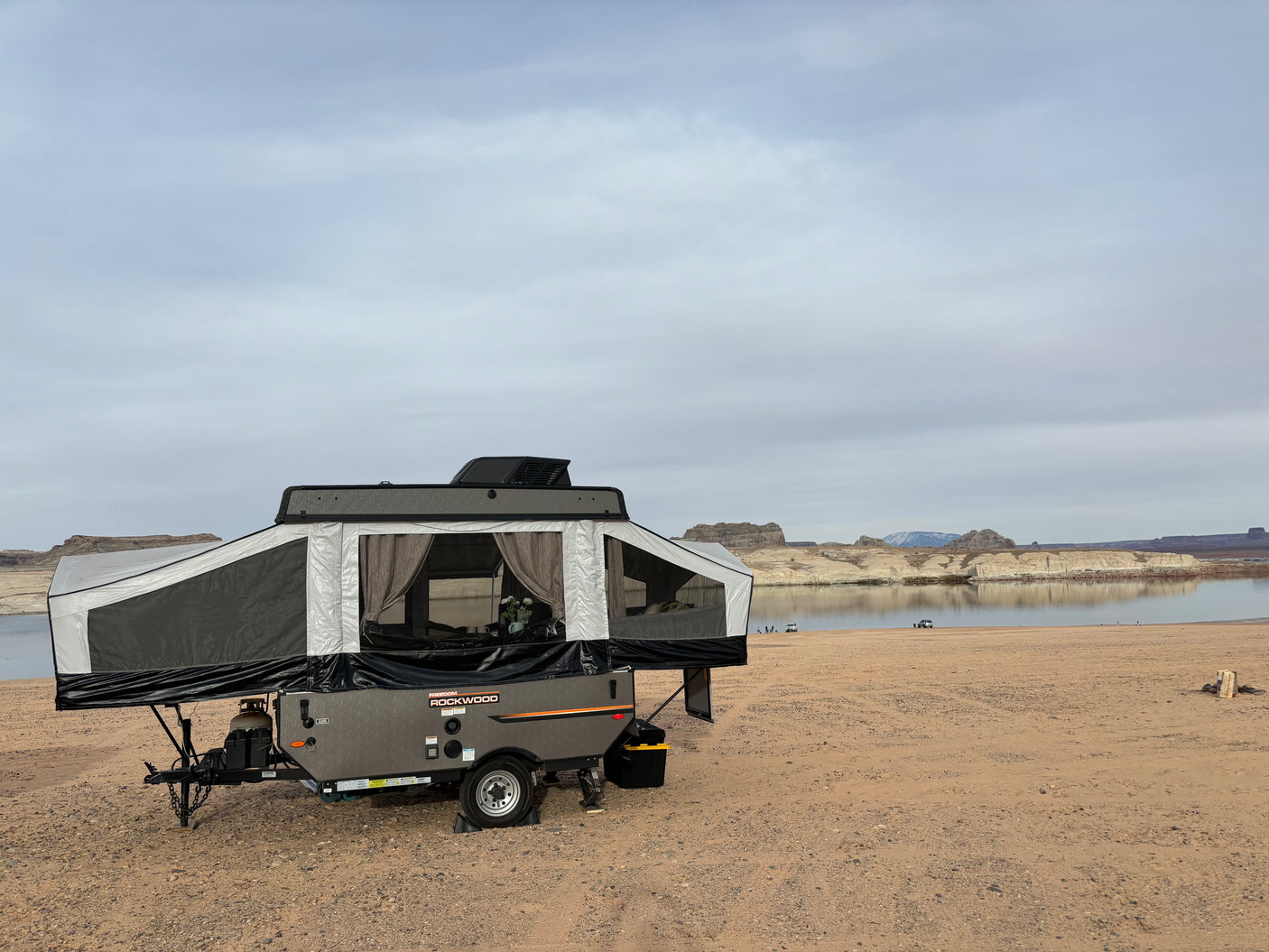 An RV rental, a grey Rockwood pop-up camper, is set up on a sandy beach next to a calm lake with cliffs beyond.