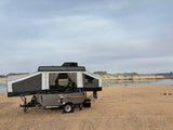 An RV rental, a grey Rockwood pop-up camper, is set up on a sandy beach next to a calm lake with cliffs beyond.