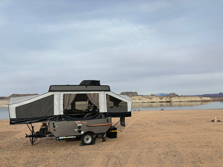 An RV rental, a grey Rockwood pop-up camper, is set up on a sandy beach next to a calm lake with cliffs beyond.
