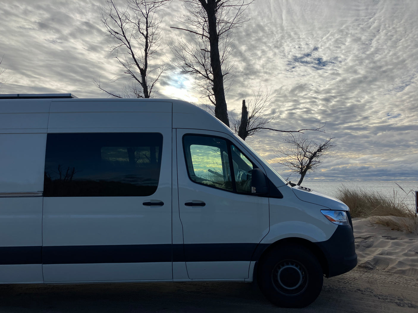 White RV rental van on a sandy beach by the water under a cloudy sky with bare trees, ready for adventure.