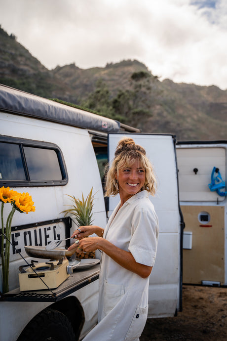Smiling woman cooking on a portable stove beside her RV rental campervan, amidst scenic mountains. Van life joy.