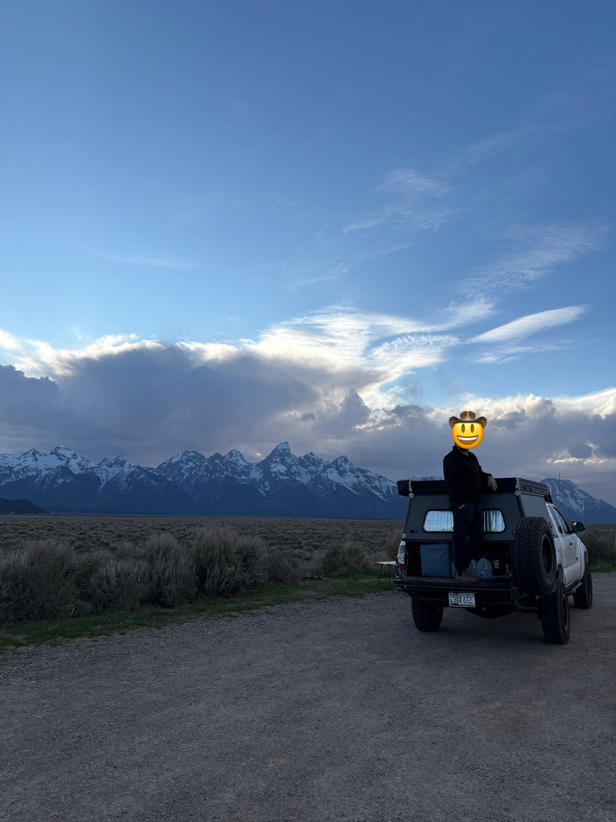 Person in a truck camper overlooking snow-capped Grand Teton mountains & vast plains. Perfect RV rental for an overland adventure!