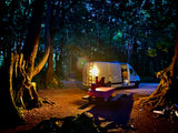 Night camping in a forest: An RV rental van is parked next to a lit picnic table, ready for a cozy evening under the trees.