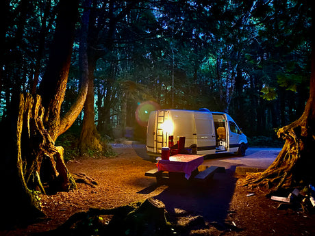 Night camping in a forest: An RV rental van is parked next to a lit picnic table, ready for a cozy evening under the trees.