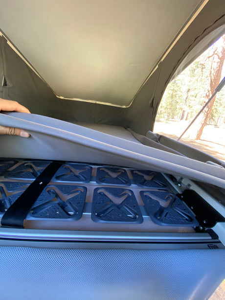 A hand lifts an RV rental mattress, revealing a Froli modular bed base for improved sleep. Pop-top tent and trees visible outside.