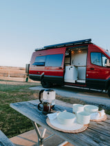 A red custom camper van parked near a picnic table with coffee and dishes, ready for your RV rental getaway.