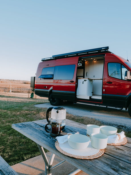 A red custom camper van parked near a picnic table with coffee and dishes, ready for your RV rental getaway.