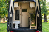 Rear view into an RV rental's compact interior: toilet on left, mini-kitchen with sink on right, and storage cabinets overhead.