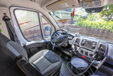 Inside a spacious RV rental van, featuring the driver's seat, dashboard, and steering wheel. Views of another RV outside.