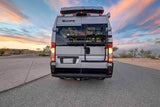 Rear view of a modern silver RV rental with a bike rack and roof box, glowing at sunset on a desert road.