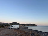 White camper van (RV rental) on a rocky beach at dawn/dusk. Calm sea and distant hills complete this serene coastal scene.