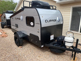 Two gray and black Clipper travel trailers, with roof racks and all-terrain tires, are parked on gravel, ideal for an RV rental.