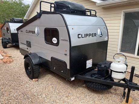 Two gray and black Clipper travel trailers, with roof racks and all-terrain tires, are parked on gravel, ideal for an RV rental.