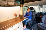 Smiling woman works on a laptop in a custom RV rental, featuring a wooden desk and sink, perfect for digital nomad travel.