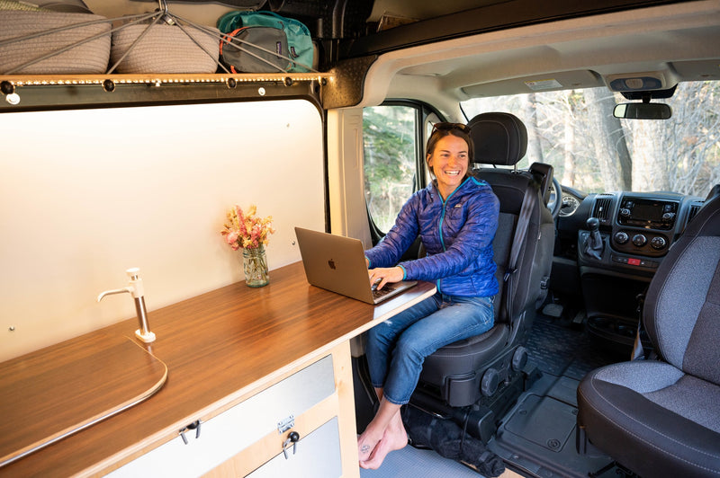 Smiling woman works on a laptop in a custom RV rental, featuring a wooden desk and sink, perfect for digital nomad travel. — photo 18