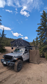 RV rental Jeep with rooftop tent & privacy shelter for off-road camping adventures under a blue sky.
