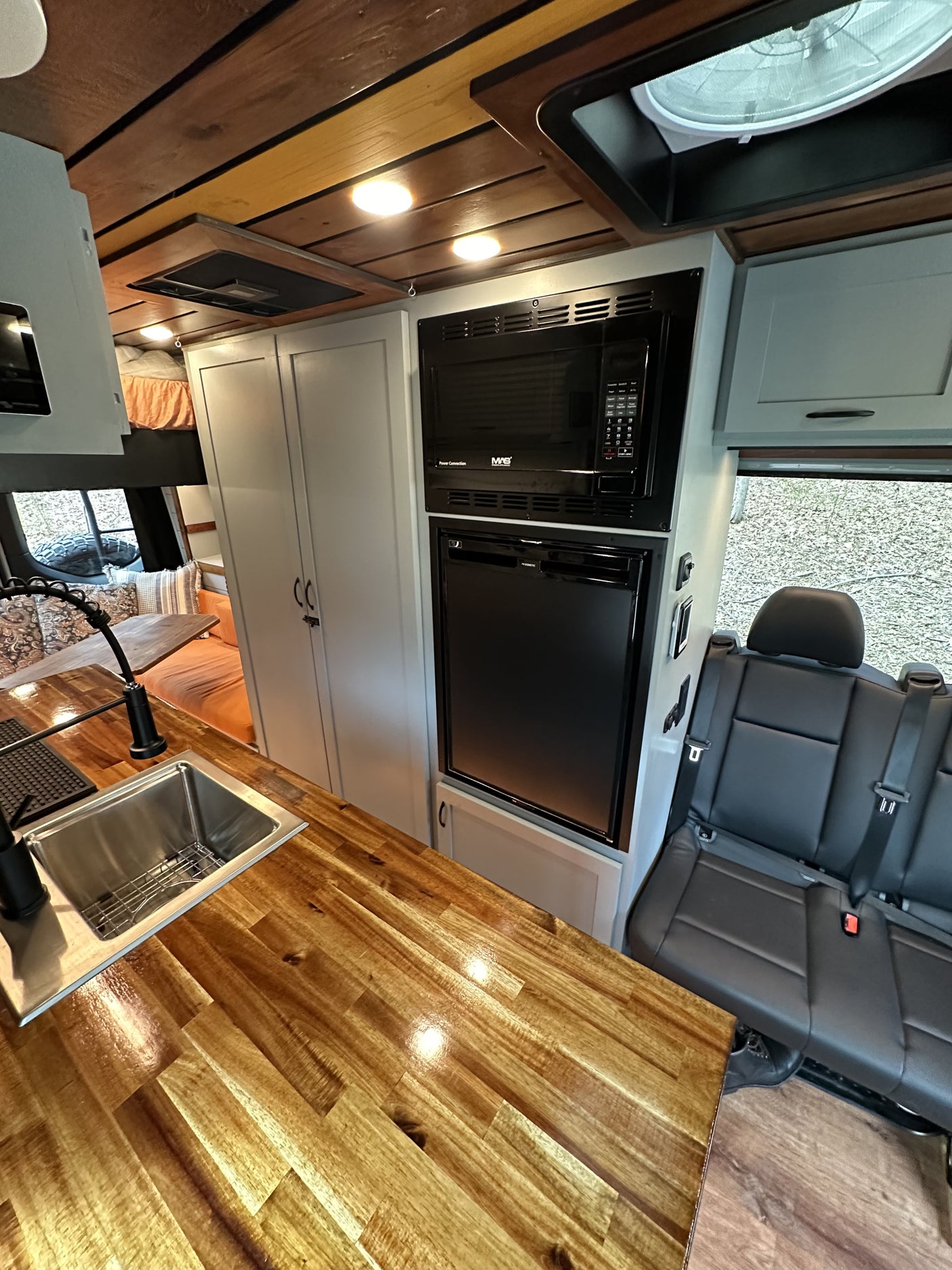 RV rental interior: sleek kitchen with glossy wood counters, sink, microwave, fridge, and black passenger seating.