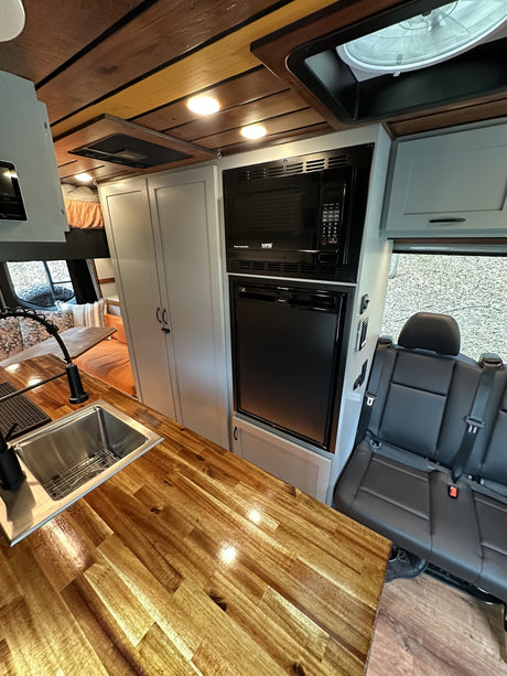RV rental interior: sleek kitchen with glossy wood counters, sink, microwave, fridge, and black passenger seating.