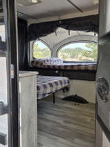 Inside an RV rental: comfy beds with plaid blankets & scenic views of nature from a pop-up camper. Ready for adventure!