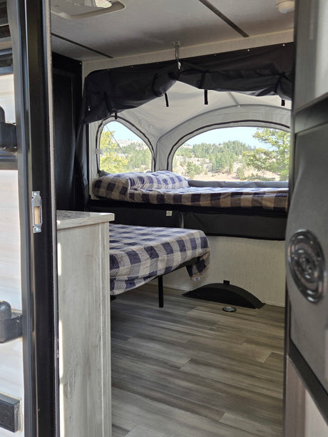 Inside an RV rental: comfy beds with plaid blankets & scenic views of nature from a pop-up camper. Ready for adventure!