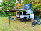RV rental: A "no bo" camper trailer with a rooftop tent, awning, chairs, and an e-bike is set up for outdoor travel.