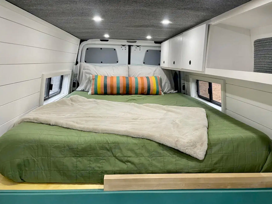 Inside this RV rental, a comfy bed with green bedding & plush blanket, white shiplap walls, and practical overhead storage.