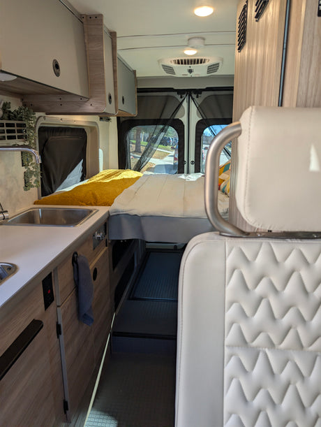 Inside an RV rental: kitchen sink, cozy bed, ceiling AC unit, and a distinctive white quilted seat in the foreground.