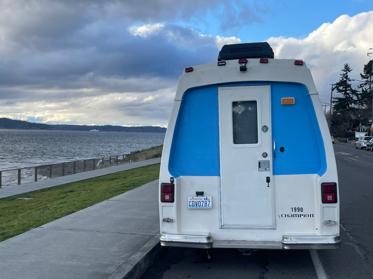 Rear of a white & blue 1990 Champion RV (possible rv rental) by a Washington waterfront, ferry in distance, cloudy sky. — photo 15