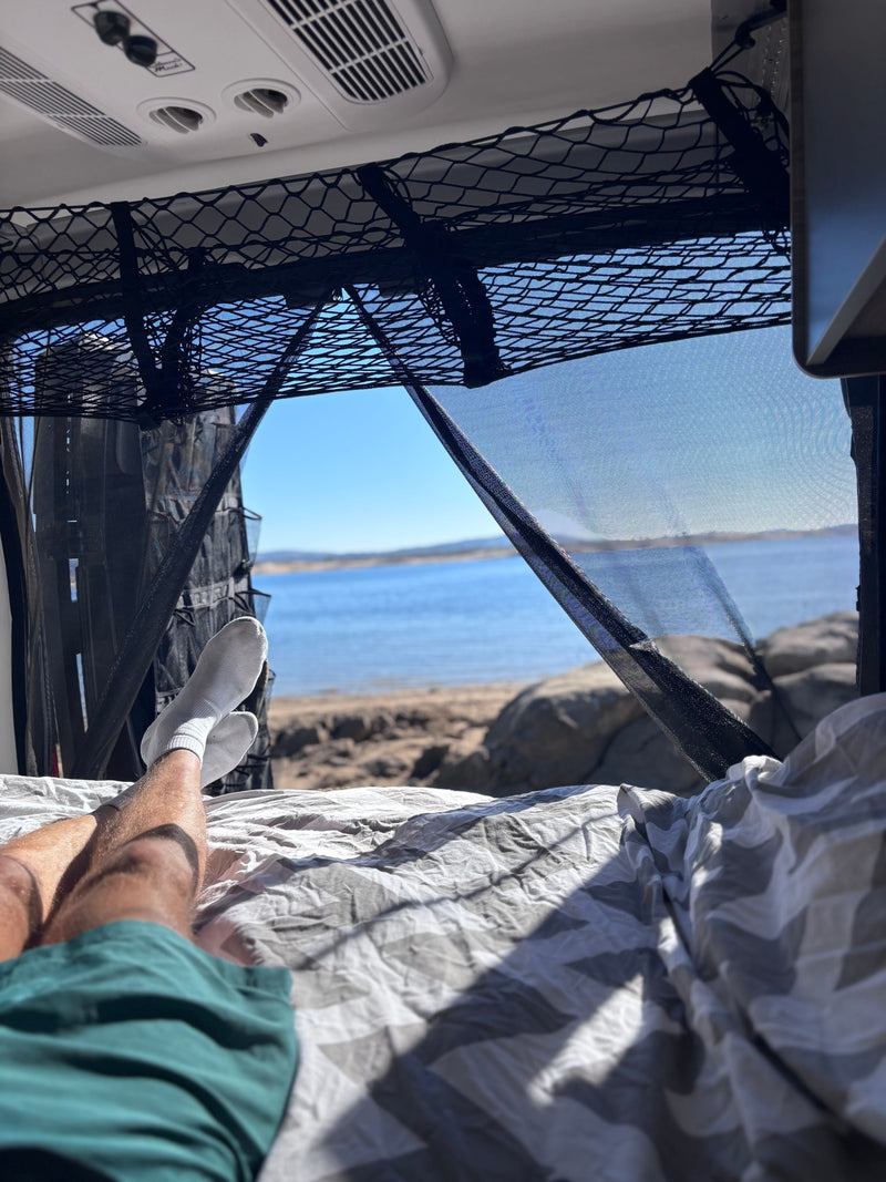 Inside an RV rental, a person's legs are stretched out on a bed, gazing at a serene lake through mesh screens. — photo 5