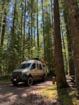 Adventure-ready RV rental in a serene forest campsite. Customized camper van under tall trees, perfect for off-road travel.