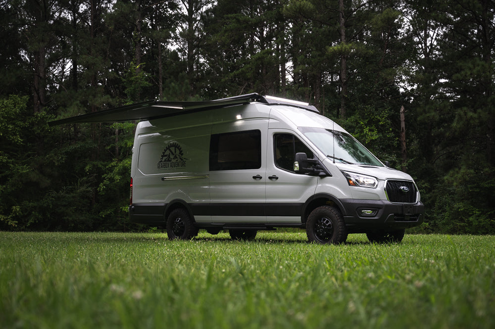 Rent this off-road adventure campervan! White Ford Transit RV rental parked in a grassy field with trees, its side awning extended.
