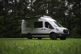 Rent this off-road adventure campervan! White Ford Transit RV rental parked in a grassy field with trees, its side awning extended.